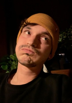 A portrait picture of Kai Klostermann. He's wearing a black sweater and a yellow beanie hat. He has a funny annoyed mimic.