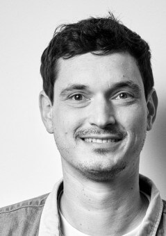 A portrait picture of Kai Klostermann. It's grayscale. He is wearing an open shirt and smirks friendly at the camera.
